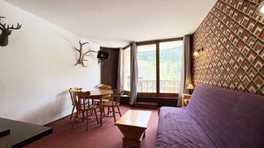 Una habitación con sofá púrpura, mesa de comedor de madera con sillas, televisor y un ventanal con vista a la vegetación.