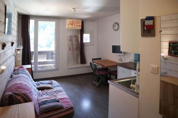 A cozy room with a bed, a dining table with chairs, a small kitchen area, and a window with curtains.