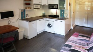 Une petite cuisine équipée d’un four à micro-ondes, d’un four et d’un lave-linge. On y trouve une petite table avec une chaise et un lit recouvert d’une couverture à rayures.