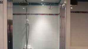 Une douche fermée par des parois en verre, dotée d’un pommeau de douche à main, d’un panneau de commande fixé au mur et d’un sol carrelé de carreaux bleus.