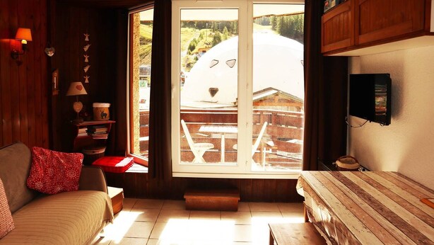 Een gezellige kamer met een bank, een houten tafel, een aan de muur bevestigde televisie en een groot raam met uitzicht op een gebouw en bomen.