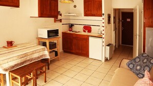 Een kleine keuken met houten kastjes, een magnetron, een koelkast, en een zithoek met een tafel en bankjes.
