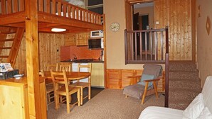 Una habitació d'estil cabana acollidora, amb taula de dinar de fusta, cadires, cuina americana, un llit individual i unes escales.
