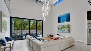 Sala de estar moderna com um sofá grande, uma mesa de centro e uma TV fixada na parede. Há uma porta de vidro deslizante que dá para uma área externa com piscina.