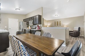 A modern kitchen with dark cabinets, a stainless steel refrigerator, and a microwave. There is a dining area with a wooden table and patterned chairs. - Short Walk to Downtown Branson & Branson Landing with Private Hot Tub & Free Tickets (Branson)