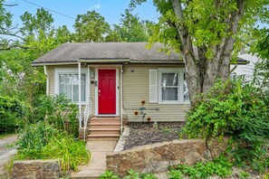 A single-story house with a red door, a small front porch, and a garden area with plants and a tree.