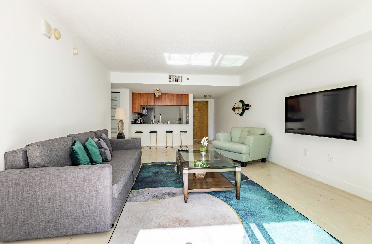 Un salon moderne avec un canapé gris, un fauteuil vert, une table basse en verre et un téléviseur à écran plat fixé au mur.