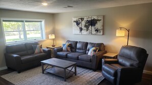 A living room with a sectional sofa, a coffee table, a leather armchair, and a floor lamp. There is a world map on the wall and a window with a view of greenery outside. - End-unit apartment with spacious lawn (Burdett)