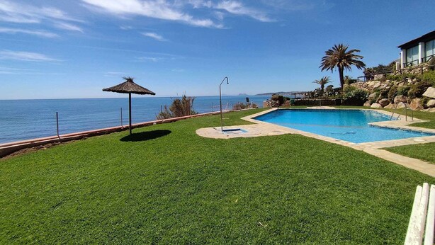 A swimming pool with a view of the ocean, a thatched umbrella, and a well-maintained lawn. - House on the beachfront. (Estepona)