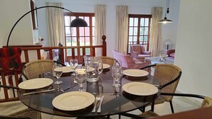 A dining area with a round table set for four, surrounded by wicker chairs. There are two windows with curtains, a sofa in the background, and a lamp above the table. - House on the beachfront. (Estepona)