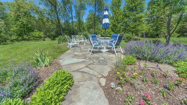 A patio with stone flooring, surrounded by lush greenery and flowering plants.