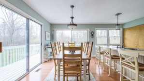 A dining area with a large table and chairs, a kitchen counter, and large windows offering a view of the outdoors.