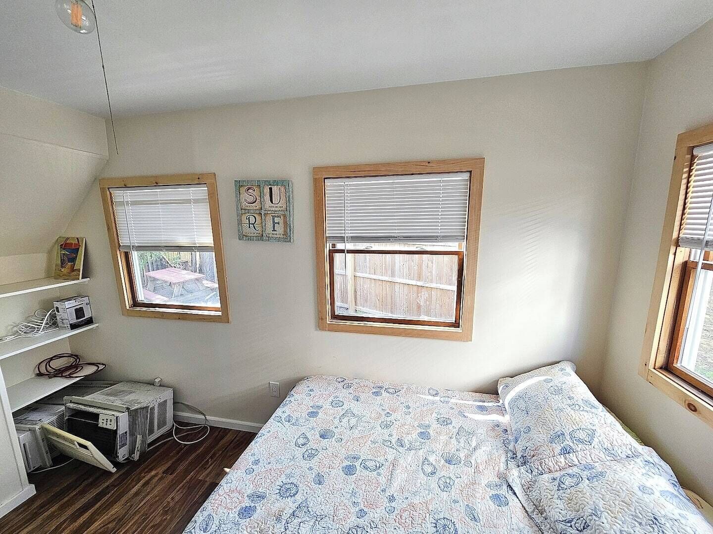 Un dormitorio con una cama, dos ventanas con persianas, un estante con objetos y una decoración en la pared.