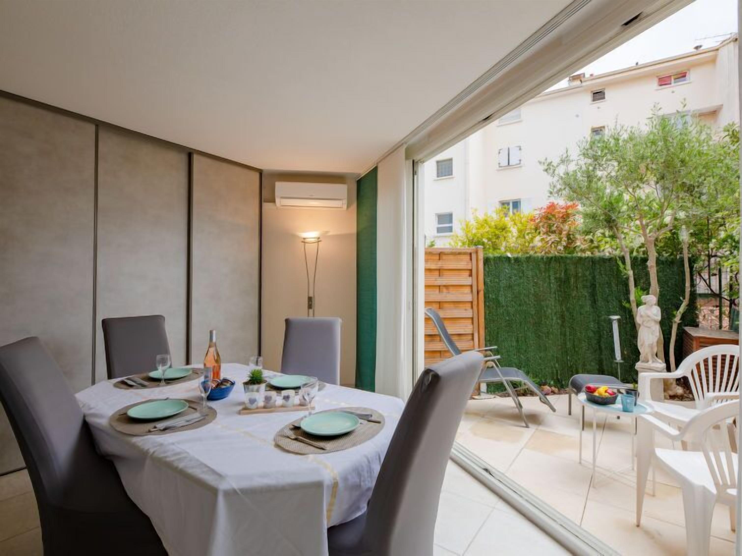 A dining area with a table set for four, a small garden visible through the glass door, and a view of a residential building in the background.