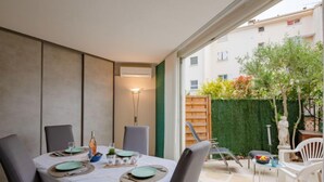 A dining area with a table set for four, a small garden visible through the glass door, and a view of a residential building in the background. - Le chant de la mer by Interhome (Fréjus)