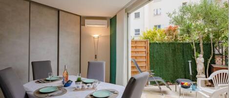 A dining area with a table set for four, a small garden visible through the glass door, and a view of a residential building in the background.