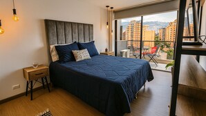A modern bedroom with a large bed, a nightstand, a small table, and a view of the city through a large window.