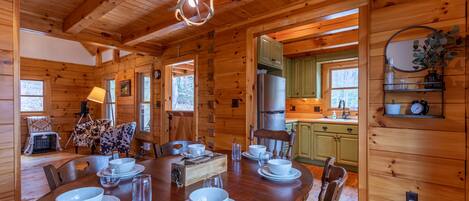 Interior de una cabaña de madera con comedor para cuatro personas, cocina con refrigerador y sala de estar con sofá y cama para perro.