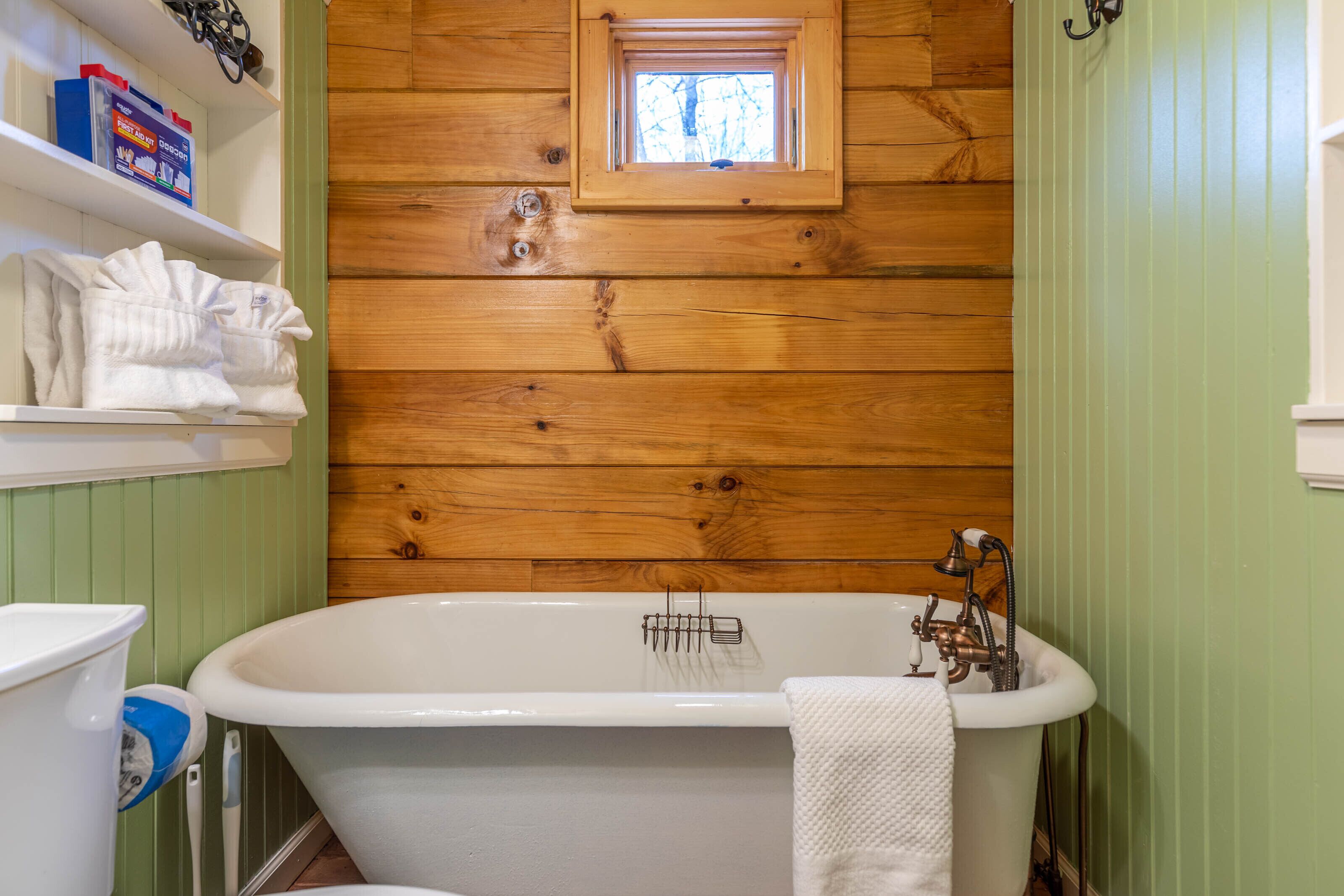 A bathroom with a freestanding bathtub, a window above the tub, and wooden paneling on the walls.