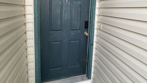 Une porte d’entrée bleu foncé avec une fenêtre au-dessus, un paillasson et un petit luminaire extérieur fixé au mur.