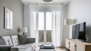 Una sala de estar moderna con un sofá gris, una mesa de centro, un televisor sobre un mueble de madera y un amplio ventanal con cortinas translúcidas.