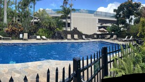 A swimming pool with lounge chairs and a stone deck, surrounded by tropical plants and a building in the background. - Beautiful Ocean view of Sunny South Shore at Poipu Beach in Kauai (Koloa)