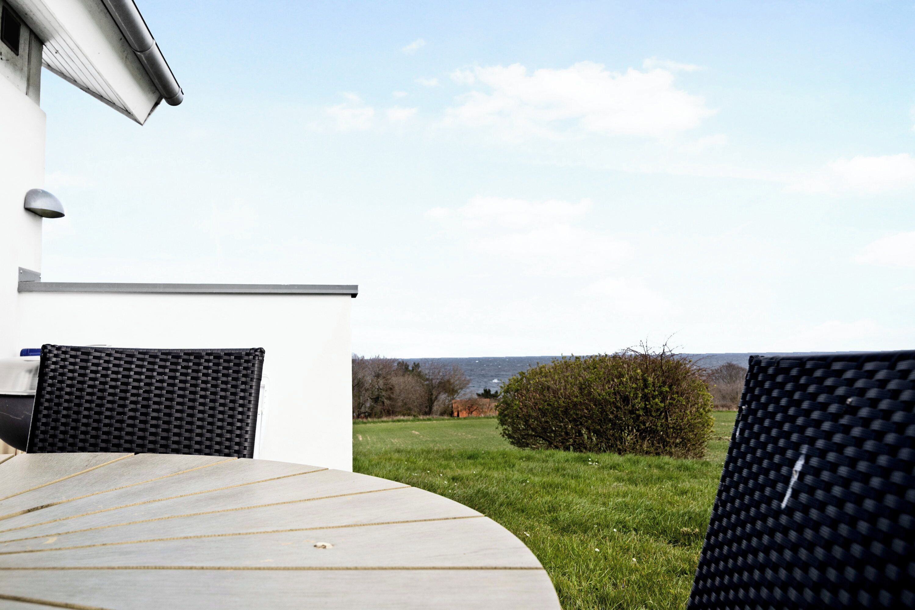 Eine Terrasse mit einem Rundtisch, einem schwarzen Stuhl und Blick auf eine Rasenfläche mit Strauch und ferne Hügel.