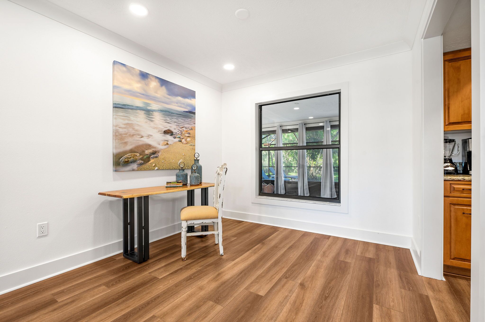 Une pièce avec un plancher en bois, un bureau avec une chaise, un grand tableau représentant une scène de plage et une fenêtre donnant sur un paysage verdoyant.