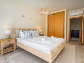 Una cama bien hecha con sábanas blancas y almohadas estampadas, una lámpara de noche, una lámpara colgante de fibra natural y un armario de madera.