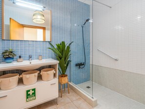 Baño con lavabo, espejo y ducha. Hay una planta y un cartel de primeros auxilios en la encimera.