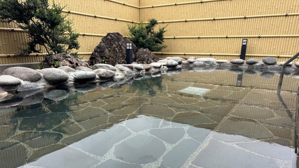 Una piscina con baldosas de piedra, un muro de bambú y rocas al fondo.
