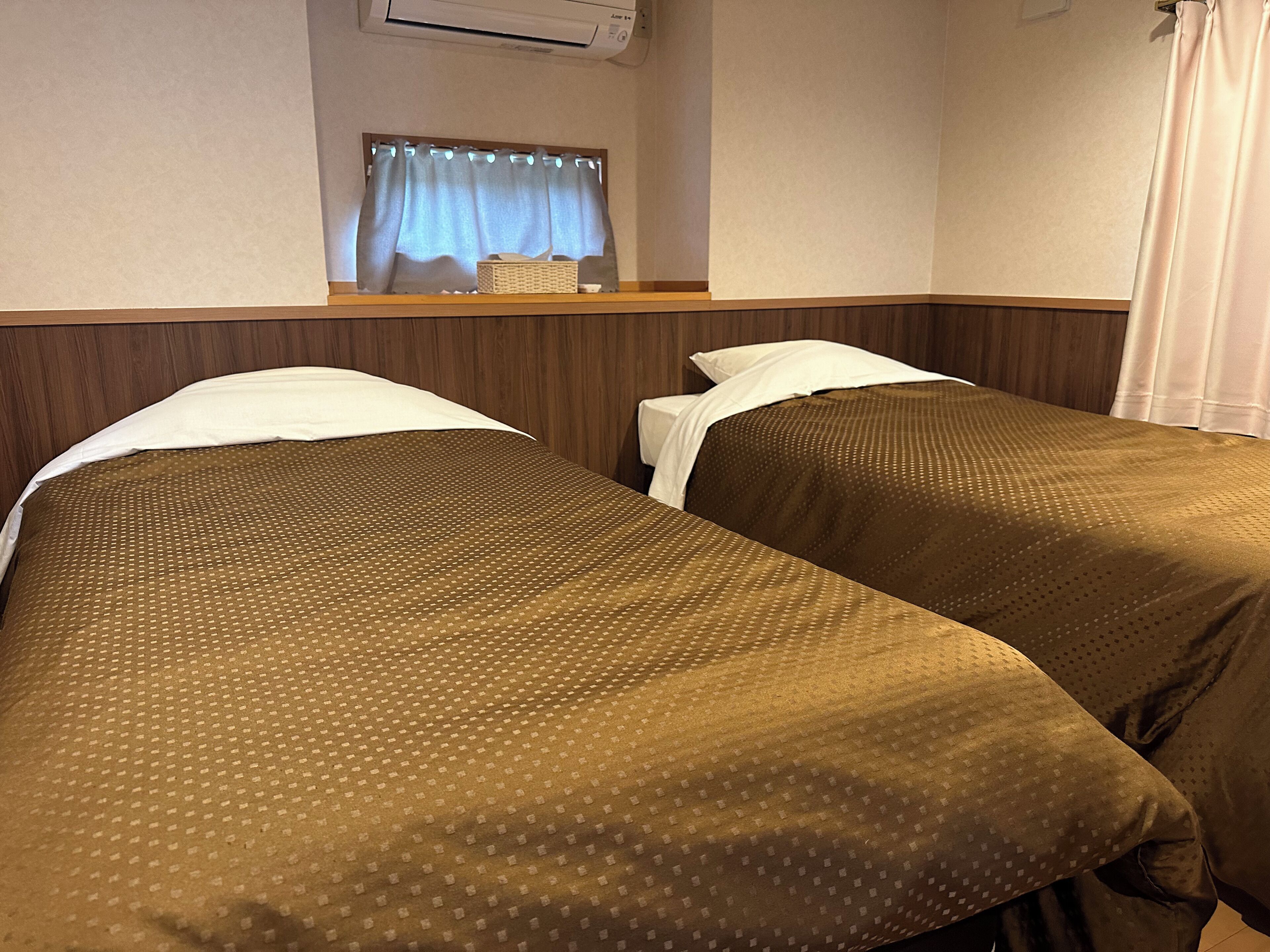 A hotel room with two single beds, a wooden headboard, a small window with curtains, and a wall-mounted air conditioner.