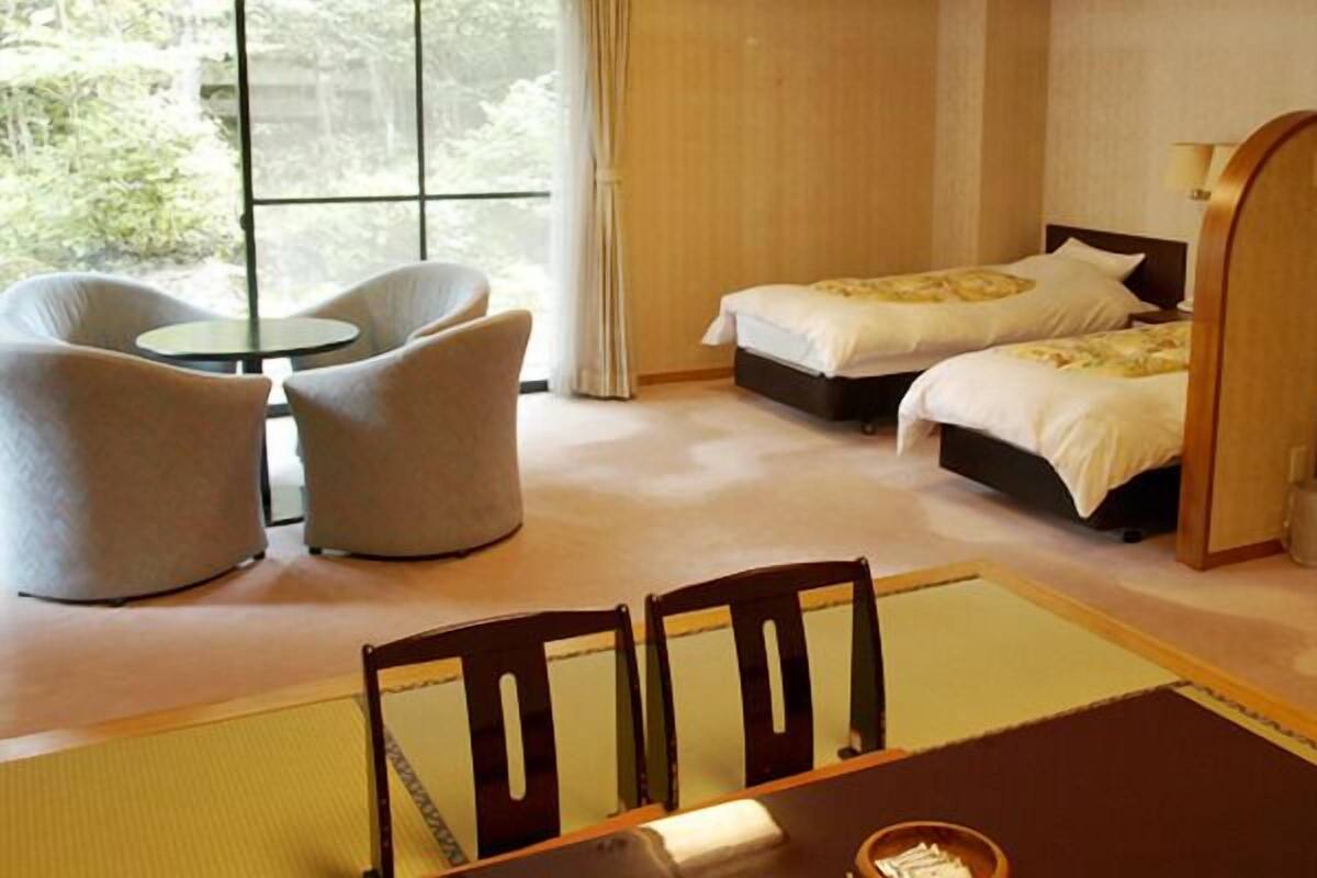 Habitación de hotel con dos camas, una mesita con sillas, una mesita de noche con lámpara y vistas a árboles a través de amplios ventanales.