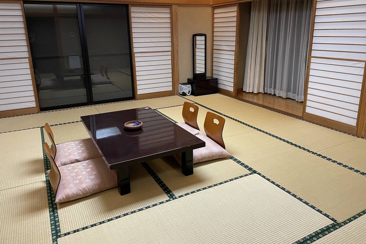 Una habitación con esterillas de tatami, una mesa baja, sillas, puertas correderas y un espejo.