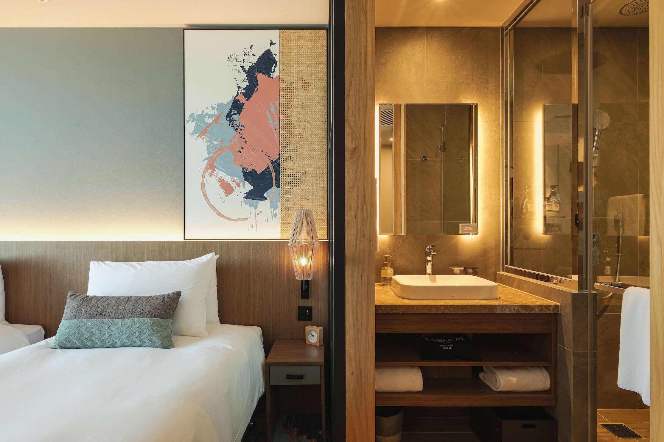 A hotel room with a bed, a nightstand, a wall-mounted clock, a large mirror, a sink, and a shower area.
