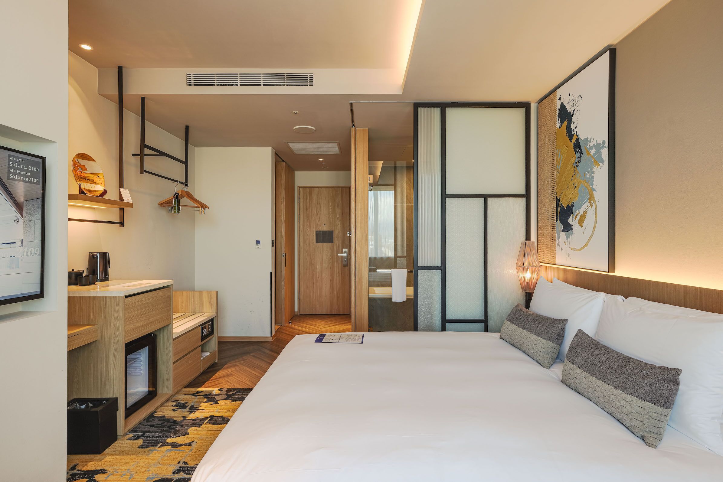 A modern hotel room with a large bed, a desk with a chair, a wardrobe, and a bathroom visible through an open door.
