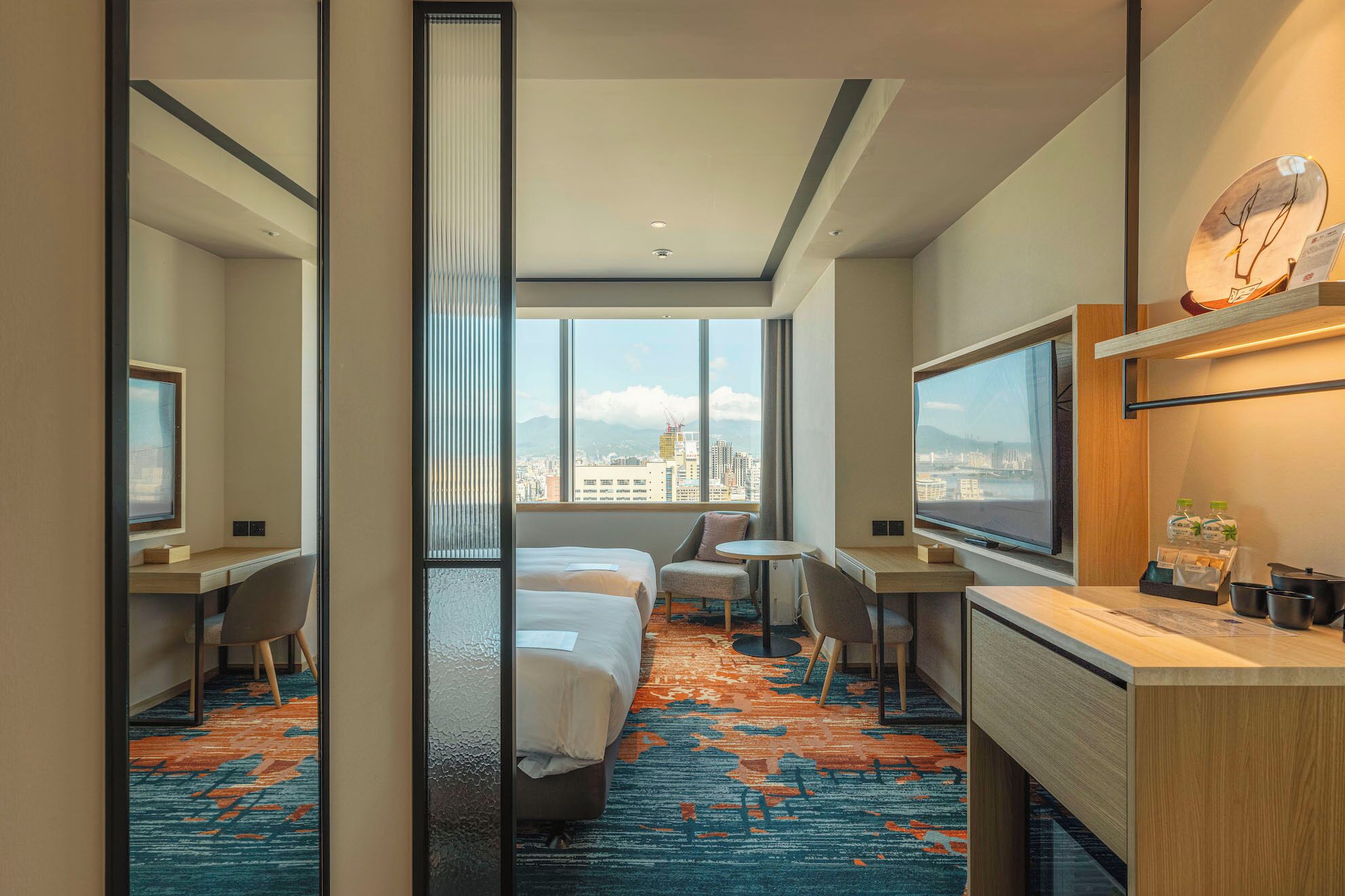 A hotel room with a bed, a desk, a chair, a TV, and a large window with a city view.