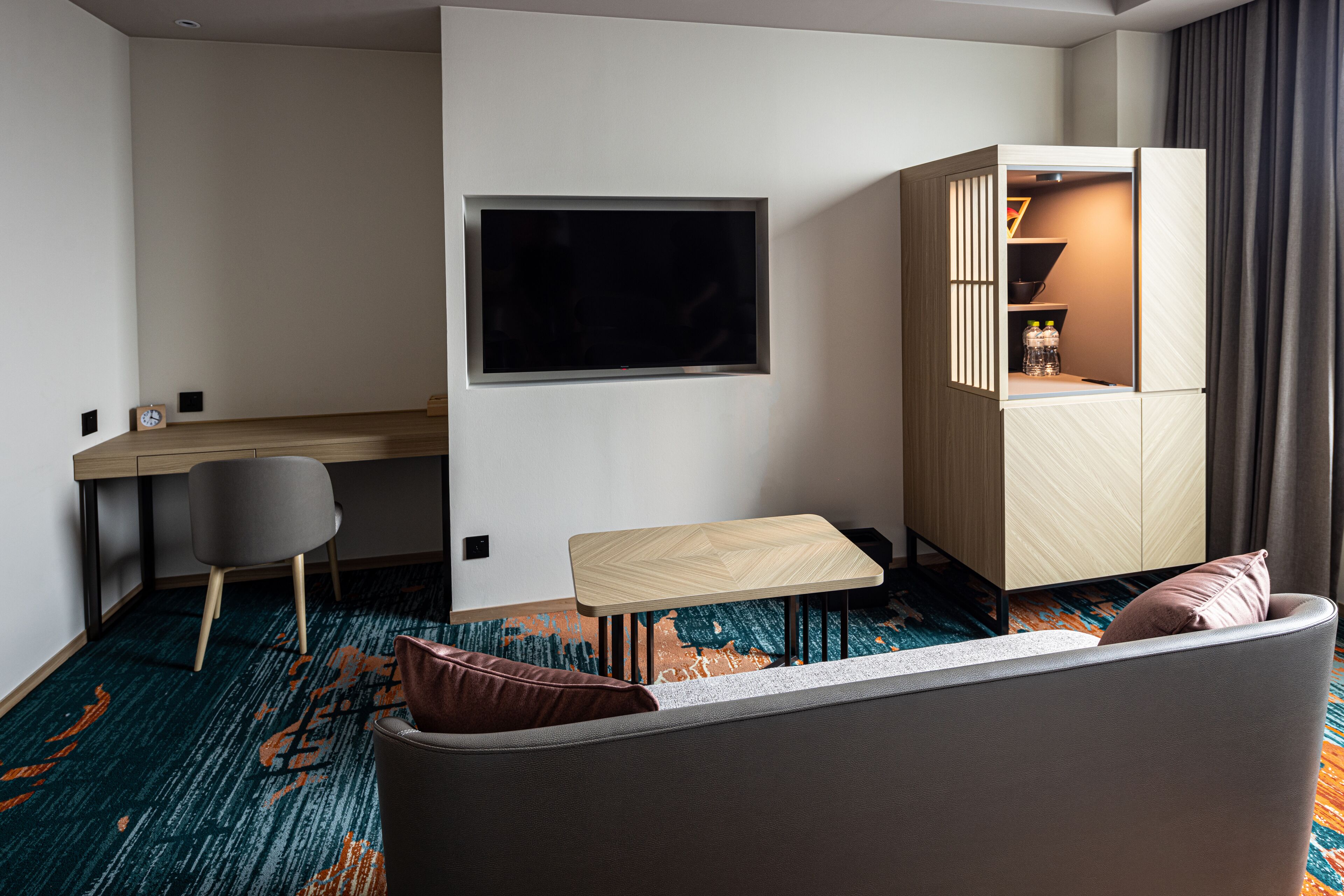 A modern hotel room with a sofa, a small table, a desk, and a TV mounted on the wall.