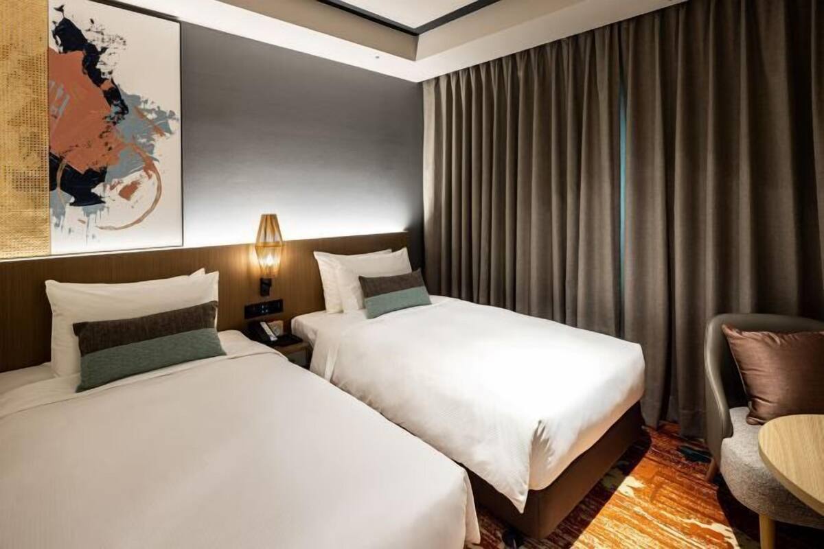 Een hotelkamer met twee bedden, een nachtkastje, een lamp, een stoel en een groot abstract schilderij aan de muur.