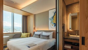 A hotel room with a large bed, a desk, a bathroom with a sink and mirror, and a view of the city.