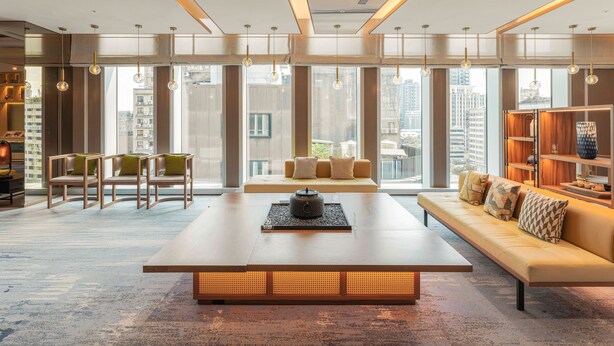 A modern living room with a large wooden coffee table, a sofa with cushions, and a bookshelf. Large windows provide a view of the cityscape.