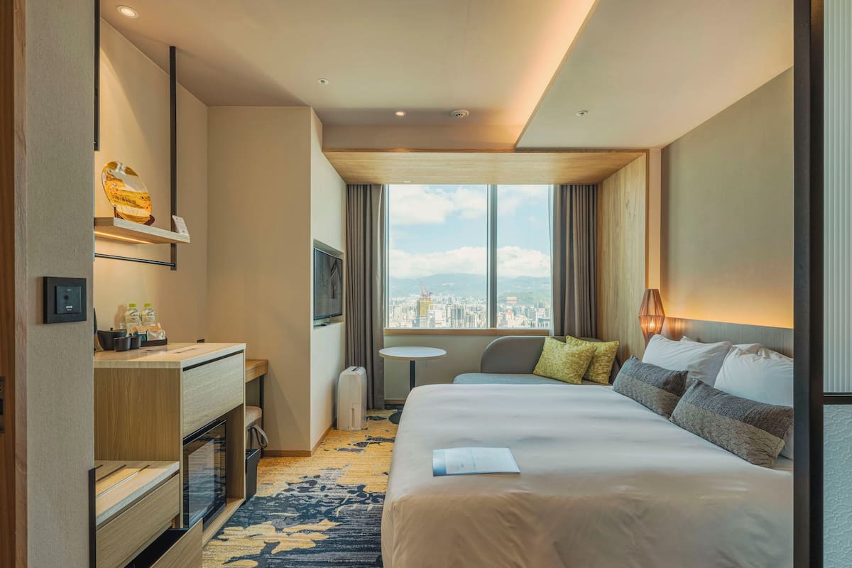 Une chambre d’hôtel comprenant un lit, un bureau, une chaise, une petite table et une grande fenêtre donnant sur la ville.