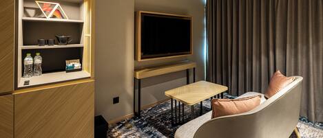 Habitación de hotel con sofá, televisor montado en la pared, mesita, armario con estantes y alfombra estampada.