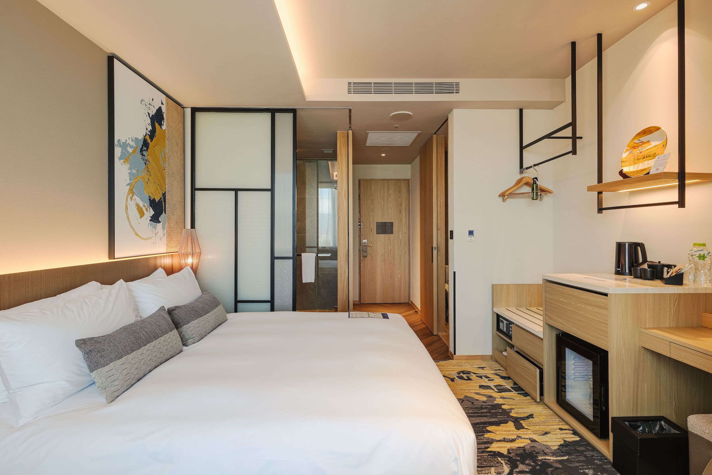 Quarto de hotel moderno com cama grande, escrivaninha de madeira, área de estar pequena e vista para o corredor.