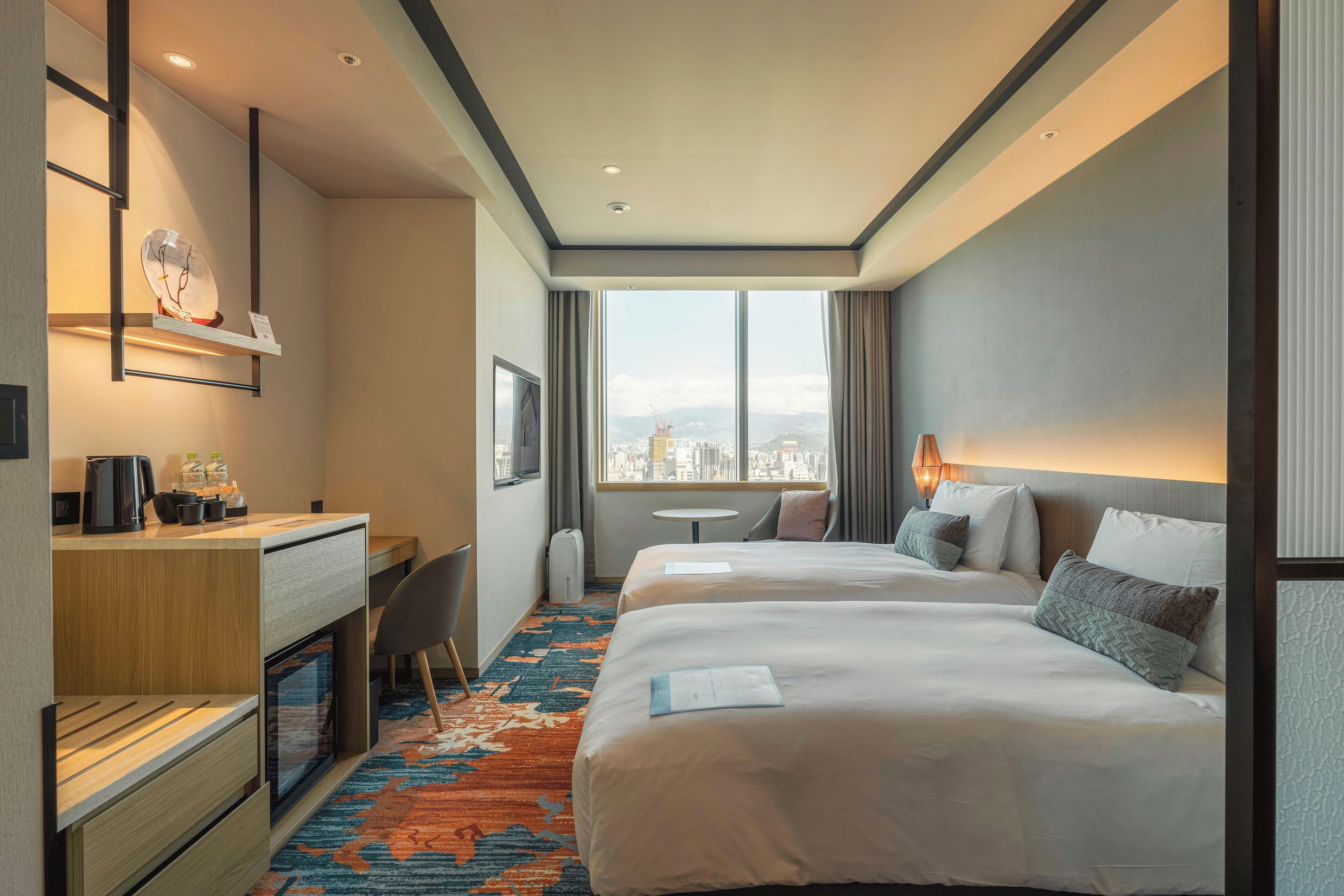 Habitación de hotel con dos camas, un escritorio, una silla, una mesita, un televisor y un ventanal con vistas a la ciudad.