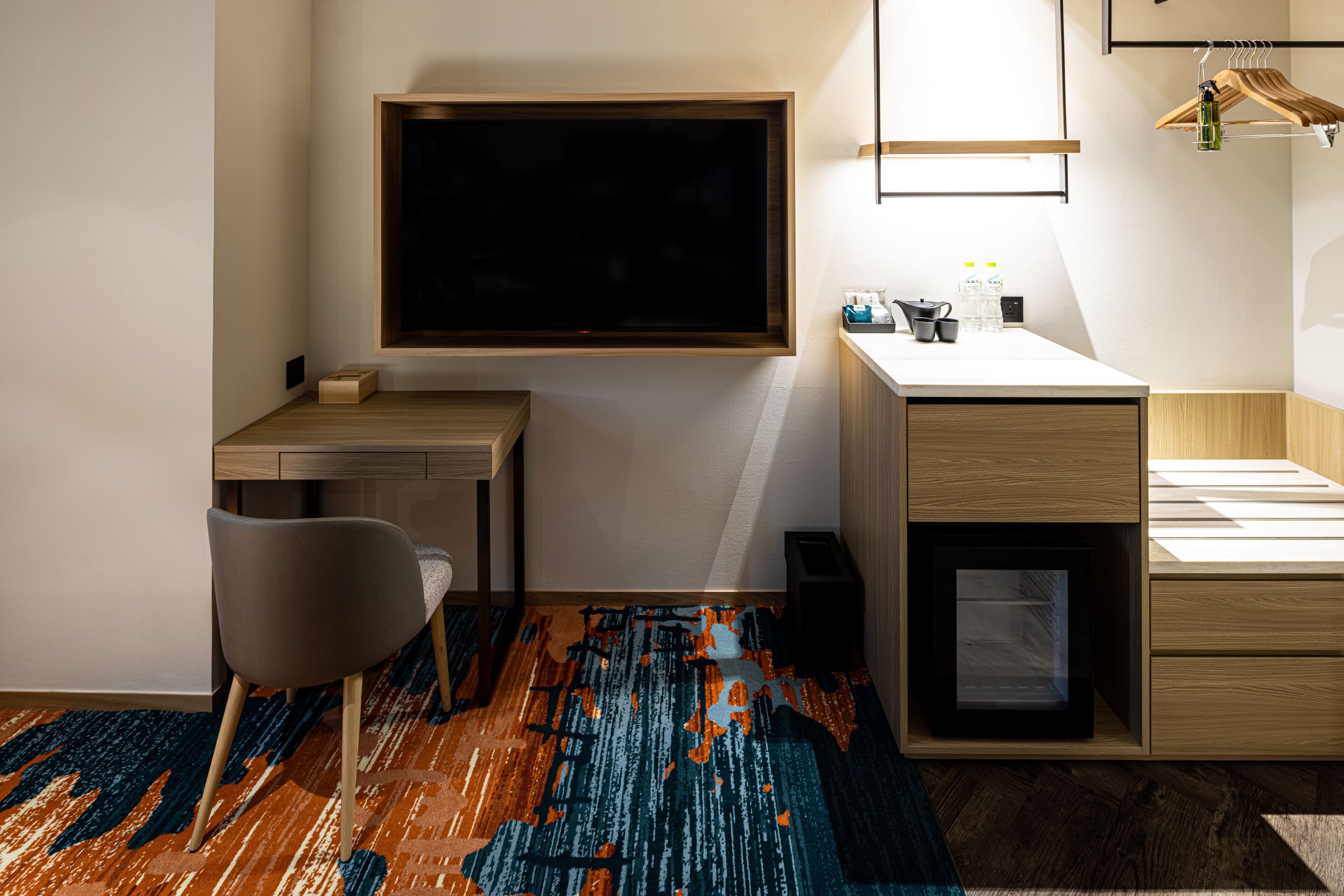 A modern room with a large TV mounted on the wall, a desk, a chair, a small cabinet, and a hanging clothes rack.