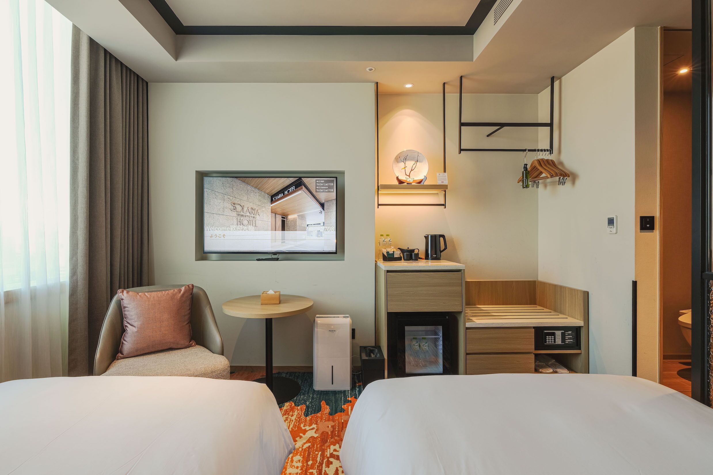 Habitación de hotel con dos camas, un escritorio, un refrigerador pequeño, una cafetera y un televisor mostrando el interior de un hotel.