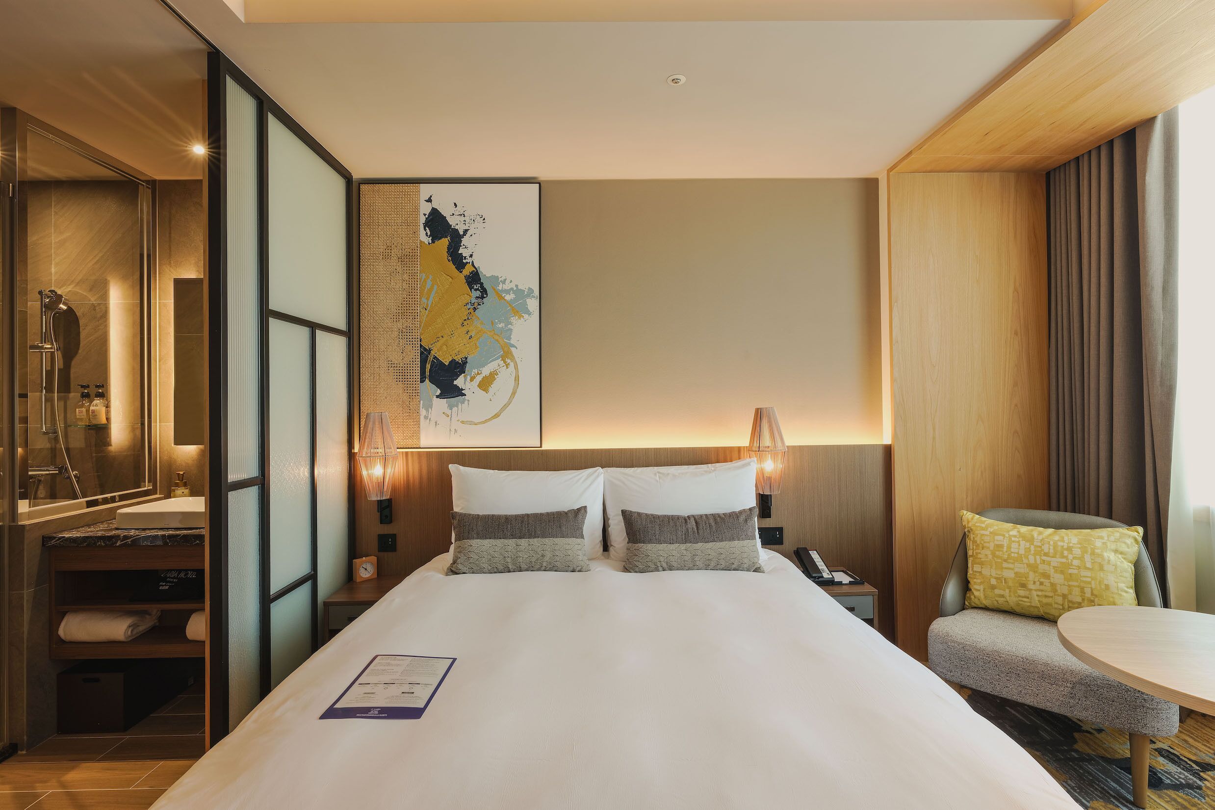 A hotel room with a large bed, a bedside table, a chair, a desk, and a large artwork on the wall.