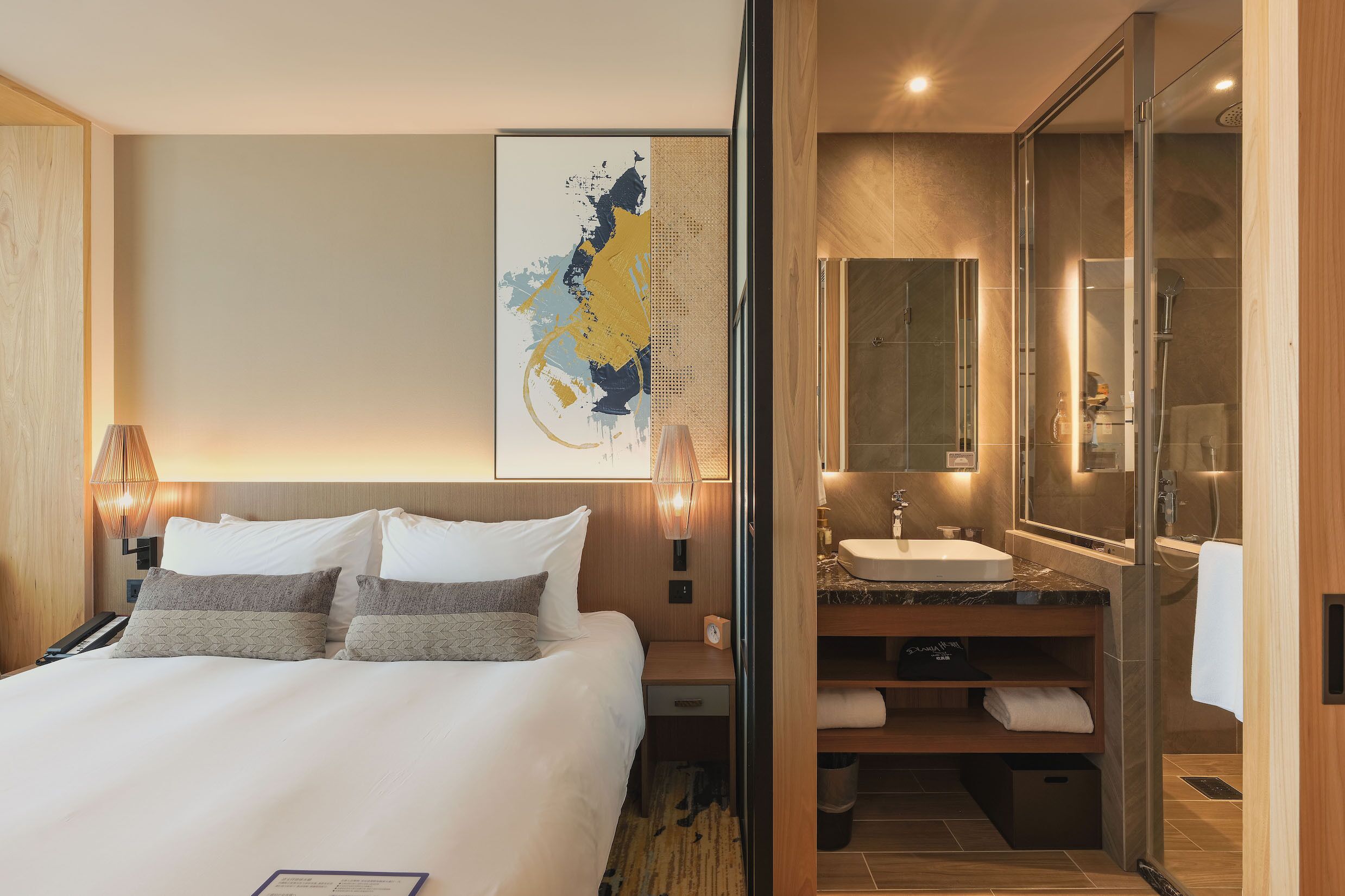 Een moderne hotelkamer met een groot bed, een nachtkastje, een wandlamp, een grote spiegel, een badkamer met wastafel en douche, en een decoratief kunstwerk aan de muur.