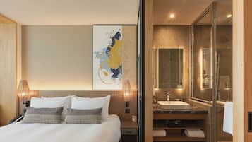 Een moderne hotelkamer met een groot bed, een nachtkastje, een wandlamp, een grote spiegel, een badkamer met wastafel en douche, en een decoratief kunstwerk aan de muur.